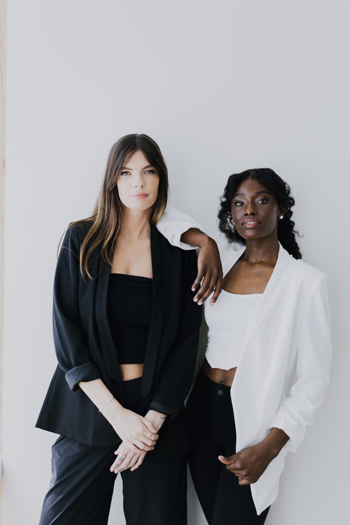 Two models in black and white blazers posed against a bright studio wall, wearing clean luminous editorial makeup