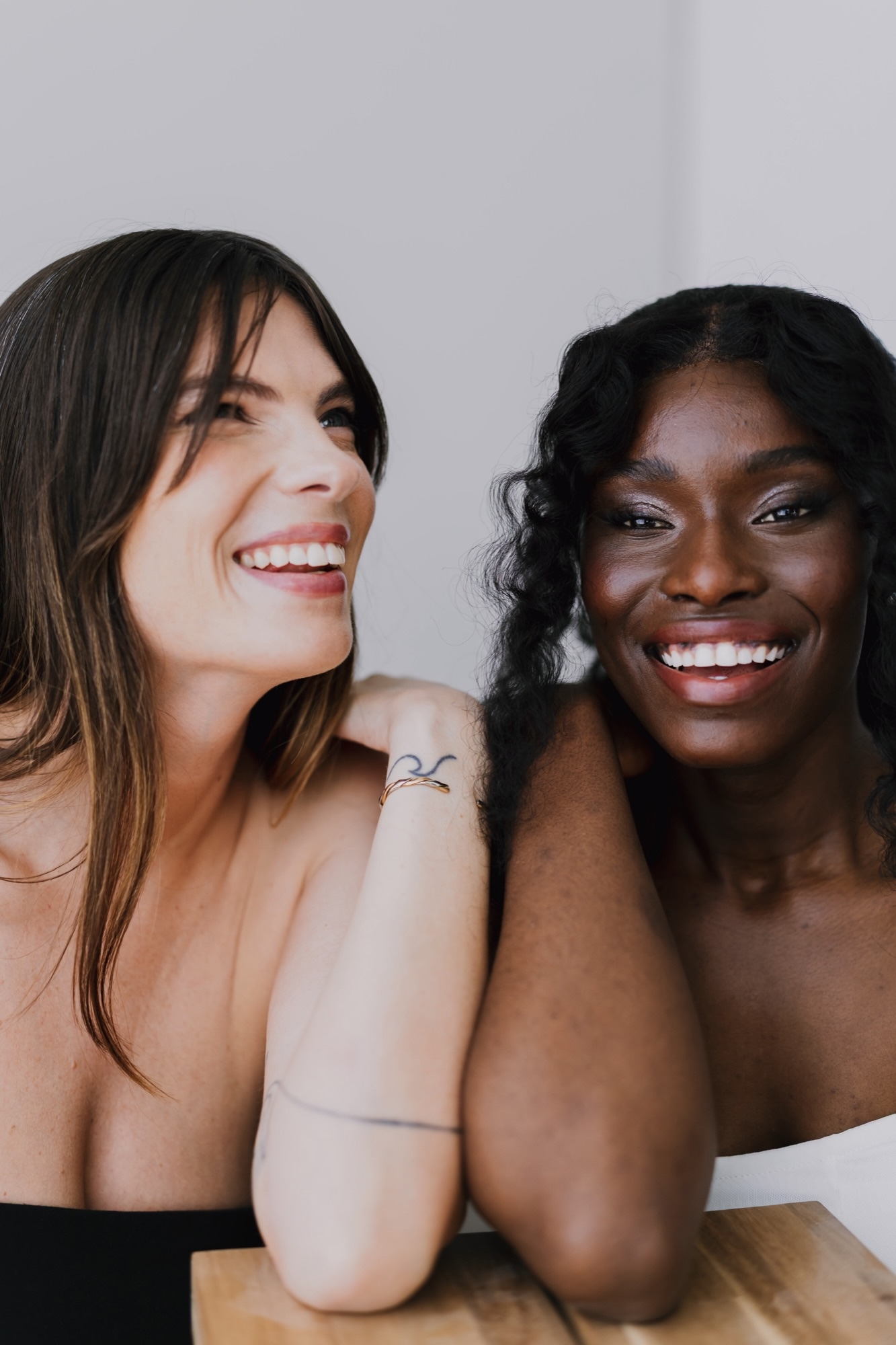 Two women smiling warmly together with soft, glowing natural makeup at a Glow Charm editorial shoot