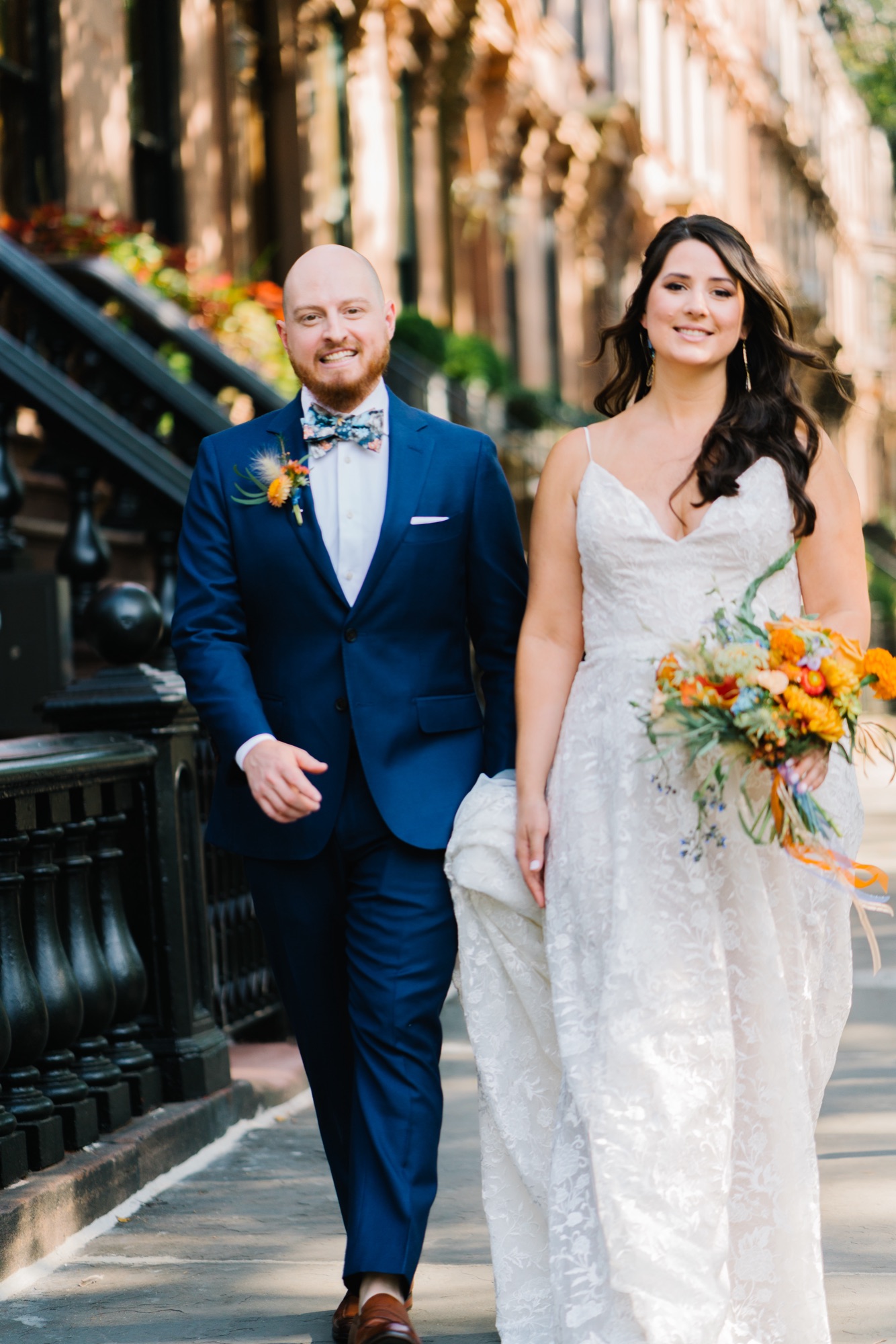 Bride and groom walking a Brooklyn brownstone sidewalk, the bride in lace with a bright orange bouquet and soft glowing makeup