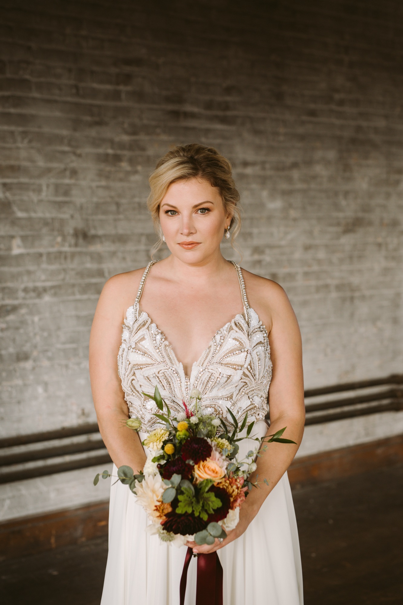 Bride in a beaded gown holding an autumn bouquet against an exposed-brick wall, natural glowing makeup